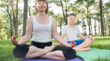 Grass Park'ta annesiyle yoga yapan genç çocuğun fotoğrafı. Aile ormanda birlikte fitness ve germe egzersizleri yapıyor