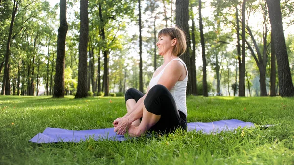 Image of mid aged smiling happy woman meditating and doing yoga exercises on grass at forest. Woman taking care of her physical and mental health while pracitising fitness and stretching at park