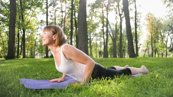 Imagem de mulher de meia-idade sorridente em roupas de fitness fazendo alongamento e exercícios de ioga. WOman meditando e fazendo esportes no tapete de fitness na grama no parque — Fotografia de Stock