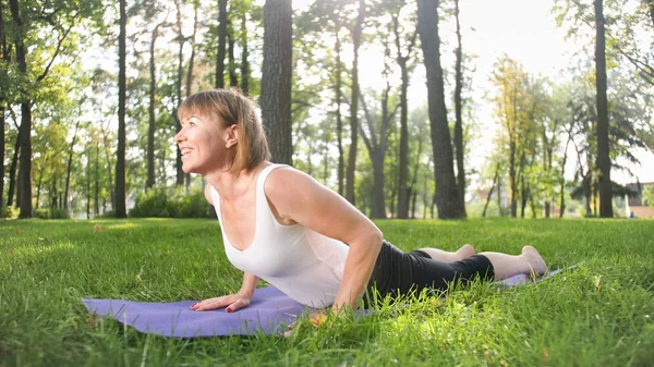 Imagem de meados da idade sorrindo mulher feliz meditando e fazendo exercícios de ioga na grama na floresta. Mulher cuidando de sua saúde física e mental enquanto pratica fitness e alongamento no parque — Fotografia de Stock