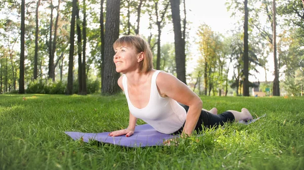 Foto de una mujer feliz sonriente de 40 años haciendo ejercicios de yoga en la alfombra de fitness en el bosque. Armonía de la naturaleza humana. Personas de mediana edad que toman coche de la salud mental y física — Foto de Stock