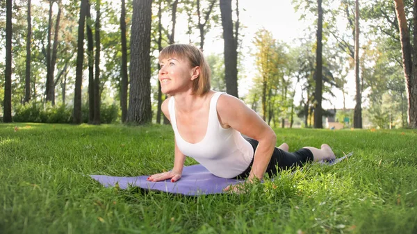 Photo of middle aged woman in sportrs clothes practicing yoga outdoors at park. Middle aged woman stretching and meditating at forest — Stock Photo, Image