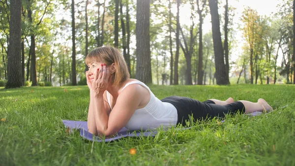 Foto de una mujer sonriente de mediana edad practicando yoga y meditando en el parque. Mujer estirando y haciendo ejercicio en la estera en el bosque — Foto de Stock