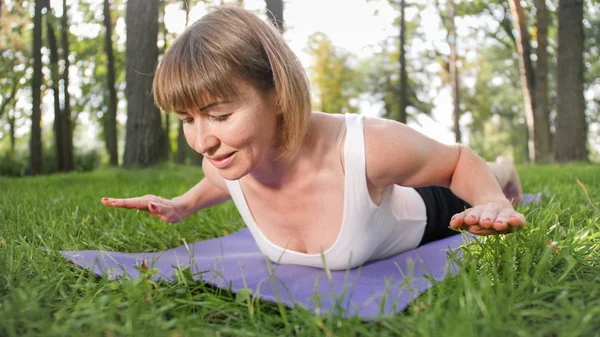 Foto de mulher sorrindo de meia-idade praticando ioga e meditando no parque. Mulher se alongando e fazendo fitness no tapete na floresta — Fotografia de Stock