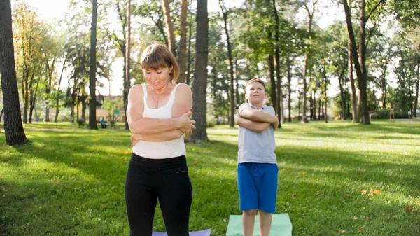 Zdjęcie nastoletniego chłopca robi jogę z matką na trawniku parku. Rodzina robi ćwiczenia fitness i rozciągania razem w lesie — Zdjęcie stockowe
