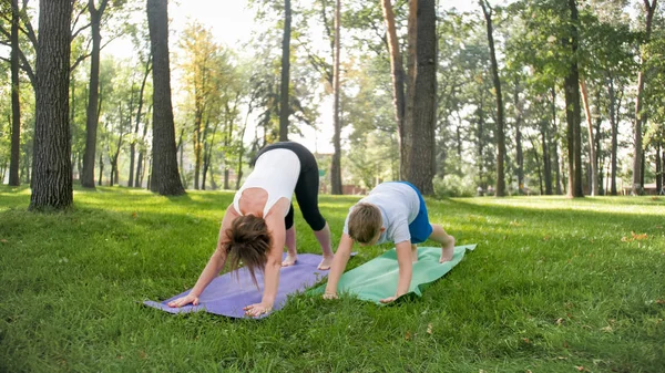 Zdjęcie nastoletniego chłopca robi jogę z matką na trawniku parku. Rodzina robi ćwiczenia fitness i rozciągania razem w lesie — Zdjęcie stockowe