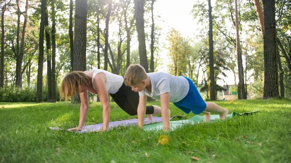 Photo of middle aged female yoga teacher or guru teaching teenage boy doing yoga. Woman with boy meditating and stretching on grass at park
