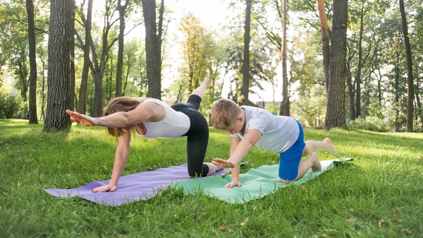 Zdjęcie nastoletniego chłopca robi jogę z matką na trawniku parku. Rodzina robi ćwiczenia fitness i rozciągania razem w lesie — Zdjęcie stockowe