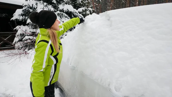 Photo of smiling girl in green coat cleaning a snow covered white car after snowfall with brush — Stock Photo, Image