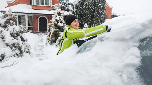 冬の雪筆を使って雪に覆われた白い車を掃除する緑のコートを着た女性の肖像画をクローズアップ — ストック写真