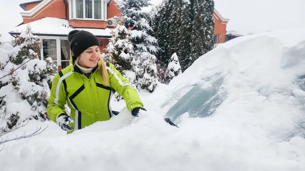 雪の嵐の後にブラシで雪に覆われた車を掃除する緑のコートで笑顔の女性のクローズアップ写真 — ストック写真