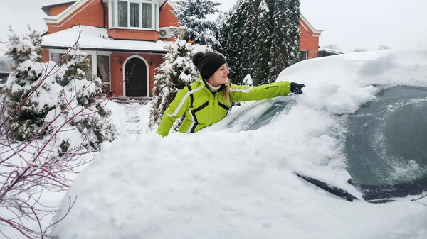 Bliska obraz uśmiechniętej Pani w zielonym płaszczu czyszczenia śniegu pokryte białym samochodem z pędzlem po burzy śnieżnej — Zdjęcie stockowe