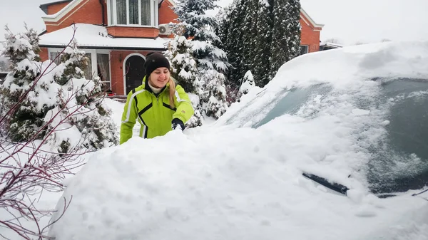 Zbliżenie strzał dziewczyny w płaszcz czyszczenia śniegu pokryte białym samochodem z pędzlem po burzy śnieżnej — Zdjęcie stockowe