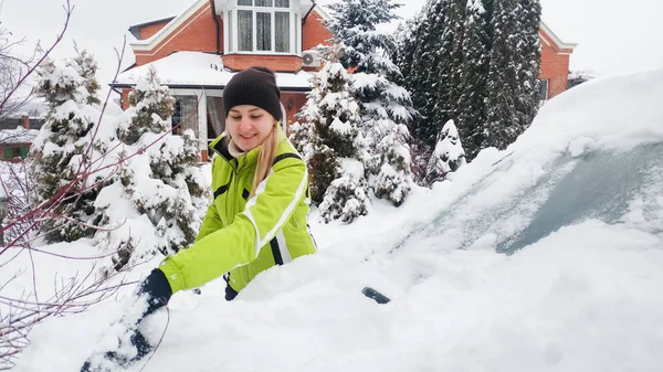 Close-up portret van glimlachend meisje in vacht schoonmaken van een sneeuw bedekt witte auto met borstel — Stockfoto