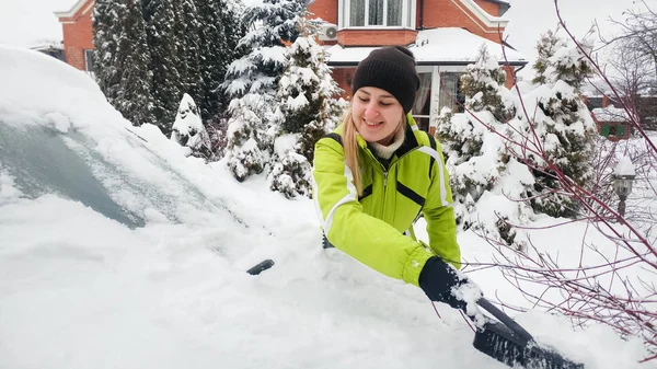 Bliska wizerunek uśmiechniętej młodej kobiety w płaszcz czyszczenia śniegu pokryte samochód po zamieć — Zdjęcie stockowe