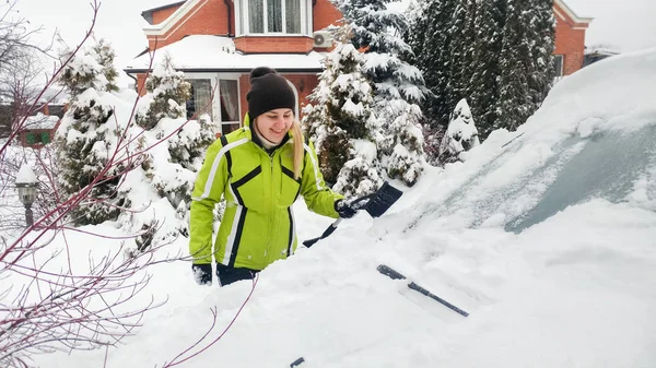 降雪後に雪に覆われた車を掃除する緑のコートを着た若い女性のクローズアップショット — ストック写真