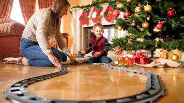 Oturma odasında Noel ağacı etrafında demiryolu inşa genç anne ile neşeli gülümseyen çocuk. Yeni Yıl veya Xmas hediye ve oyuncak alan çocuk