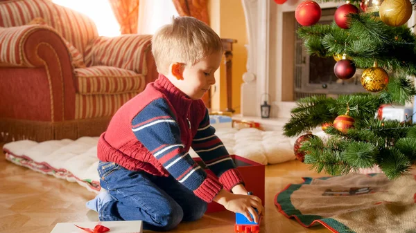 Porträt eines fröhlich lächelnden Jungen, der Spielzeug aus einer Weihnachtsgeschenkschachtel nimmt und auf dem Boden unter dem Weihnachtsbaum spielt — Stockfoto