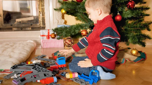 Malý 4-letý chlapec, který sedí na podlaze pod vánočním stromku a staví železnici z hraček — Stock fotografie