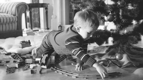 Preto e branco imagem de 4 anos de idade menino brincando com brinquedo ferroviário e trem no chão na sala de estar sob a árvore de Natal — Fotografia de Stock