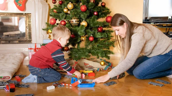 Retrato de niño sonriente feliz con la madre construyendo el ferrocarril y jugando con el tren de juguete en el suelo bajo el hermoso árbol de Navidad —  Fotos de Stock