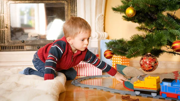 Retrato de menino sentado no chão na sala de estar e olhando para o trem de brinquedo em ferrovias sob a árvore de Natal — Fotografia de Stock