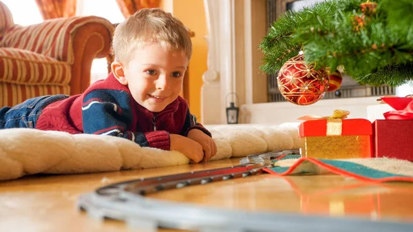 Feliz niño riendo tumbado en el suelo y mirando el ferrocarril de juguete alrededor de un gran árbol de Navidad —  Fotos de Stock