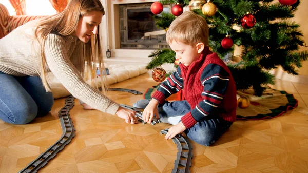Madre joven ayudando a su hijo pequeño a construir sendas para el tren de juguete bajo el árbol de Navidad en la sala de estar. Niño recibiendo regalos y juguetes en Año Nuevo o Navidad —  Fotos de Stock