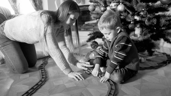 Черно-белый образ матери и маленького мальчика, играющих и строящих игрушечную железную дорогу в гостиной в рождественское утро. Дети получают подарки и игрушки на Новый год или Рождество — стоковое фото