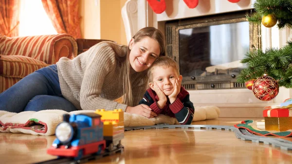 Retrato de la feliz familia sonriente tendida en el suelo y mirando el tren cabalgando en el ferrocarril bajo el árbol de Navidad en la sala de estar —  Fotos de Stock