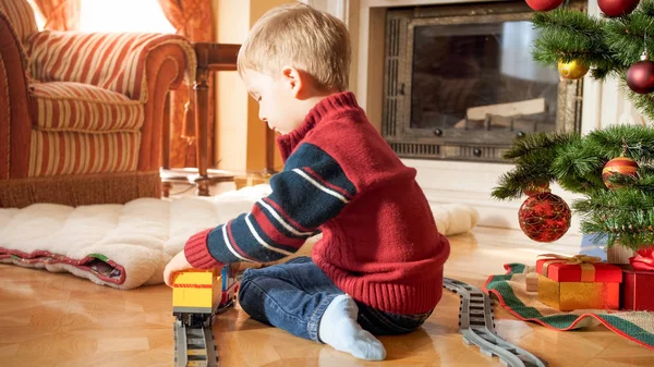 Porträt eines kleinen dreijährigen Jungen, der neben dem Weihnachtsbaum sitzt und mit einer Spielzeugeisenbahn spielt. Kind erhält an Neujahr oder Weihnachten Geschenke und Spielzeug — Stockfoto