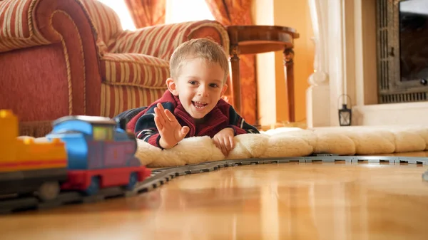 Porträtt av glad leende liten pojke som ligger på golvet och viftande hand till leksakståg ridning på järnväg i vardagsrummet. — Stockfoto