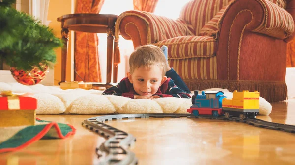 Getöntes Bild eines lächelnden kleinen Jungen, der auf Holzboden mit seiner neuen Spielzeugeisenbahn und Eisenbahn spielt. Kind erhält an Neujahr oder Weihnachten Geschenke und Spielzeug — Stockfoto