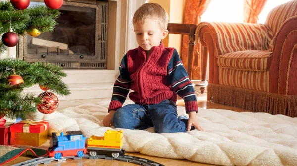 Retrato de menino sentado no chão e brincando com o brinquedo ferroviário que o Papai Noel lhe deu para o Natal. Criança recebendo presentes e brinquedos no Ano Novo ou Natal — Fotografia de Stock