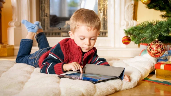 Retrato de close-up de menino deitado no chão na sala de estar decorado para o Natal e jogando no computador tablet digital — Fotografia de Stock