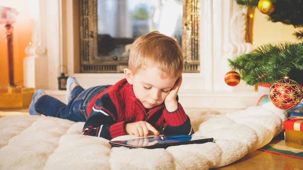 Getönte Nahaufnahme Porträt eines kleinen Jungen, der unter dem Weihnachtsbaum auf weichem Teppich liegt und auf einem digitalen Tablet-Computer spielt — Stockfoto