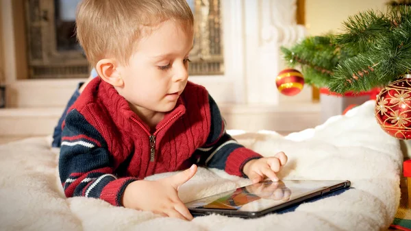 Oturma odasında Noel ağacının altında yerde yatarken dijital tablet bilgisayar kullanarak mutlu gülümseyen küçük çocuk Closeup portre — Stok fotoğraf
