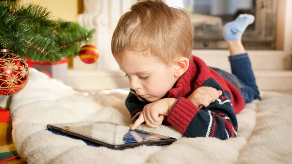 Noel ağacının yanında battaniyenin üzerinde yatan ve dijital tablet bilgisayarda oynayan küçük çocuğun portresi — Stok fotoğraf