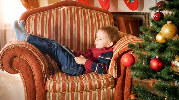 Retrato de menino deitado em poltrona ao lado da bela árvore de Natal decorada e jogando no computador tablet digital — Fotografia de Stock