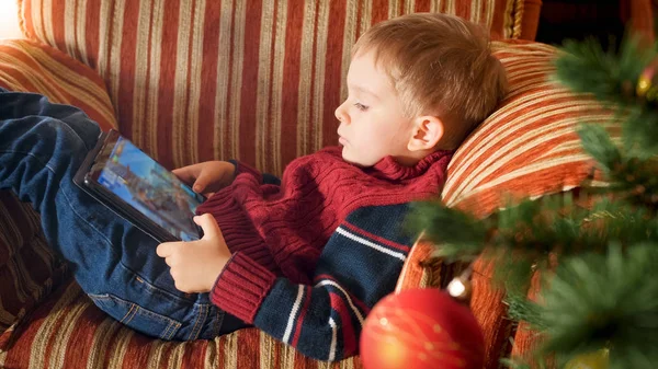 Porträt eines kleinen Jungen mit digitalem Tablet-Computer, der im Sessel neben dem Weihnachtsbaum sitzt — Stockfoto