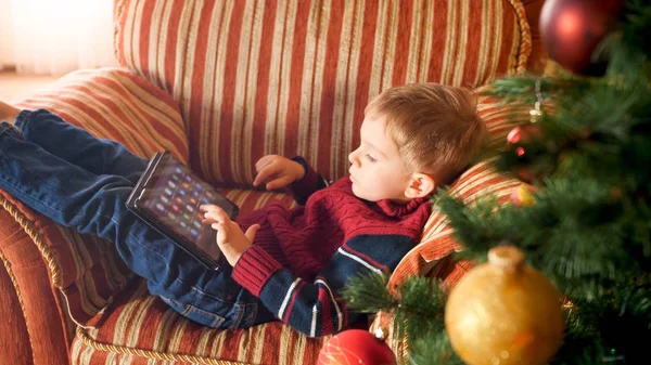 Portret małego chłopca z cyfrowym tabletem komputerowym grając w gry lub przeglądając Internet. Dziecko w domu na Boże Narodzenie rano — Zdjęcie stockowe