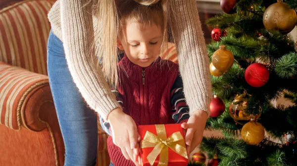 Foto einer jungen Mutter, die an Weihnachten Surpirse macht und ihrem kleinen Sohn eine Geschenkbox neben dem Weihnachtsbaum im Wohnzimmer schenkt — Stockfoto