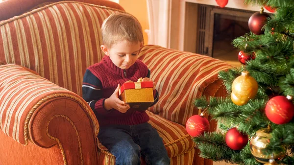 Porträt eines kleinen Jungen, der im Sessel sitzt und eine Weihnachtsgeschenkschachtel vom Weihnachtsmann in der Hand hält — Stockfoto