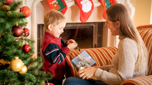 リビングルームでクリスマスの朝にプレゼントやプレゼントを包む小さな男の子と笑う若い母親の肖像画 — ストック写真