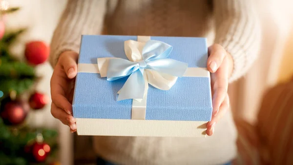 Foto di primo piano di giovane donna in piedi all'albero di Natale e dando scatola regalo blu con fiocco nastro di seta. Immagine perfetta per vacanze invernali e celebrazioni — Foto Stock