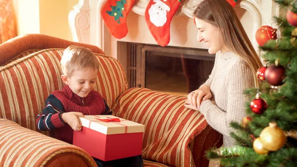 Портрет счастливой матери, делающей рождественский подарок своему маленькому сыну, сидящему в кресле рядом с красивой елкой. Идеальный образ для зимних праздников и торжеств — стоковое фото
