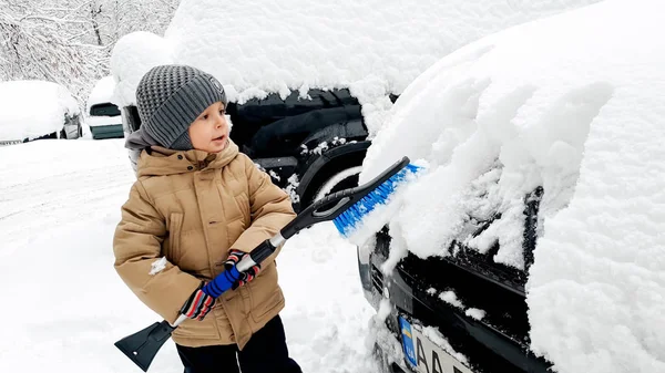 Foto de cerca del adorable niño en abrigo beige que ayuda a limpiar el coche cubierto de nieve con cepillo azul —  Fotos de Stock