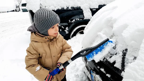 Zbliżenie strzał adorable szczęśliwy chłopiec w płaszcz pomaga oczyścić śnieg pokryte samochód z pędzlem — Zdjęcie stockowe