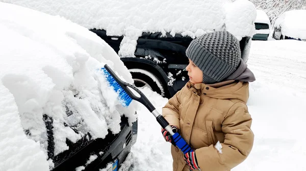 Закройте фотографию очаровательного мальчика в бежевом пальто, вычищающего машину после снежной бури кистью — стоковое фото
