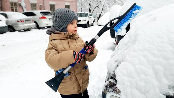 Nahaufnahme eines Jungen im beigen Mantel, der das Auto nach einem Schneesturm mit einem Pinsel säubert — Stockfoto